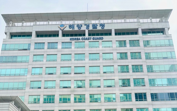 Four individuals are posing for a group photo in front of the Korea Coast Guard (해양경찰청) headquarters building. The group includes one person in U.S. Army multicam/OCP camouflage uniform, one civilian in a white shirt with a lanyard/badge, and two individuals in dark navy blue uniforms consistent with U.S. Coast Guard attire. The building is a large modern multi-story glass and stone structure with both Korean and English signage.