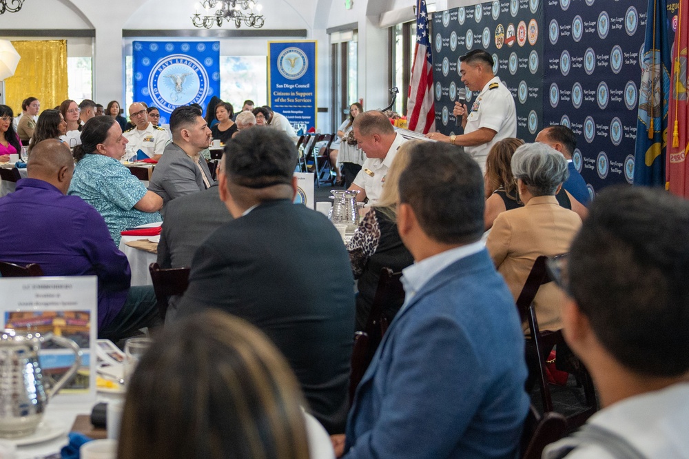 A formal Navy League San Diego Council event, likely an awards luncheon or recognition ceremony, is taking place in a banquet hall. A naval officer in dress whites is speaking at a podium or standing at the front of the room while attendees seated at round tables listen. Banners and backdrops for the Navy League San Diego 'Citizens in Support of the Sea Services' are prominently displayed.