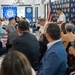 A formal Navy League San Diego Council event, likely an awards luncheon or recognition ceremony, is taking place in a banquet hall. A naval officer in dress whites is speaking at a podium or standing at the front of the room while attendees seated at round tables listen. Banners and backdrops for the Navy League San Diego 'Citizens in Support of the Sea Services' are prominently displayed.