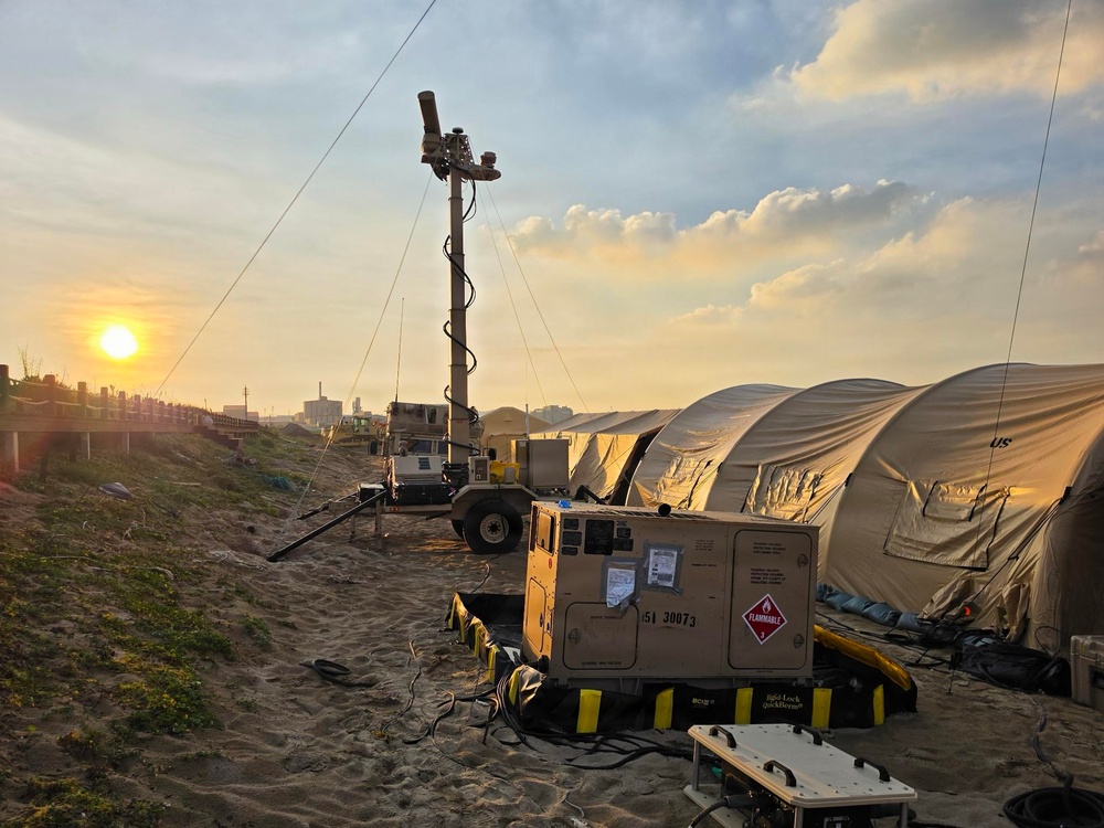 A military forward operating base or field camp is set up on sandy terrain at sunset or sunrise. The scene includes military-grade equipment including a generator, surveillance/lighting mast, tactical vehicles, and large tan military shelter tents. The setup suggests an active field operation or humanitarian mission deployment.