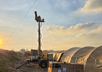 A military forward operating base or field camp is set up on sandy terrain at sunset or sunrise. The scene includes military-grade equipment including a generator, surveillance/lighting mast, tactical vehicles, and large tan military shelter tents. The setup suggests an active field operation or humanitarian mission deployment.