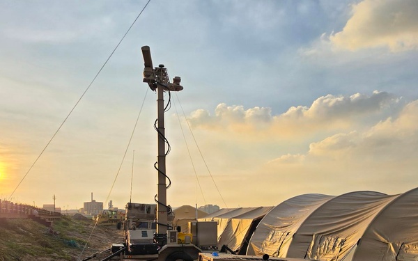 A military forward operating base or field camp is set up on sandy terrain at sunset or sunrise. The scene includes military-grade equipment including a generator, surveillance/lighting mast, tactical vehicles, and large tan military shelter tents. The setup suggests an active field operation or humanitarian mission deployment.