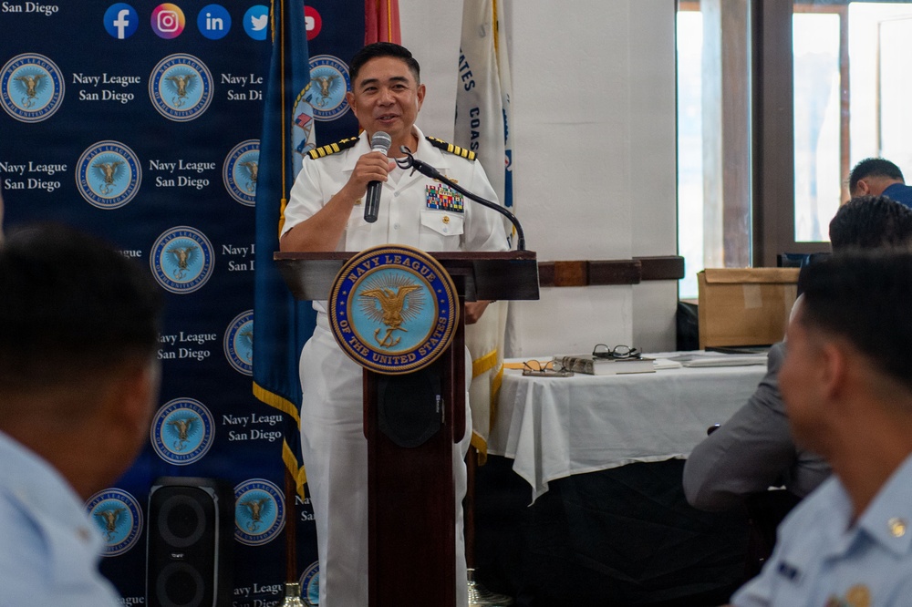 A senior U.S. Navy officer in white dress uniform is delivering a speech at a podium bearing the Navy League of the United States seal, at a Navy League San Diego event. The officer displays multiple rows of service ribbons and gold sleeve stripes indicating a high rank. An audience is partially visible in the foreground, and flags including a U.S. Coast Guard flag are visible in the background.