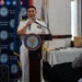 A senior U.S. Navy officer in white dress uniform is delivering a speech at a podium bearing the Navy League of the United States seal, at a Navy League San Diego event. The officer displays multiple rows of service ribbons and gold sleeve stripes indicating a high rank. An audience is partially visible in the foreground, and flags including a U.S. Coast Guard flag are visible in the background.
