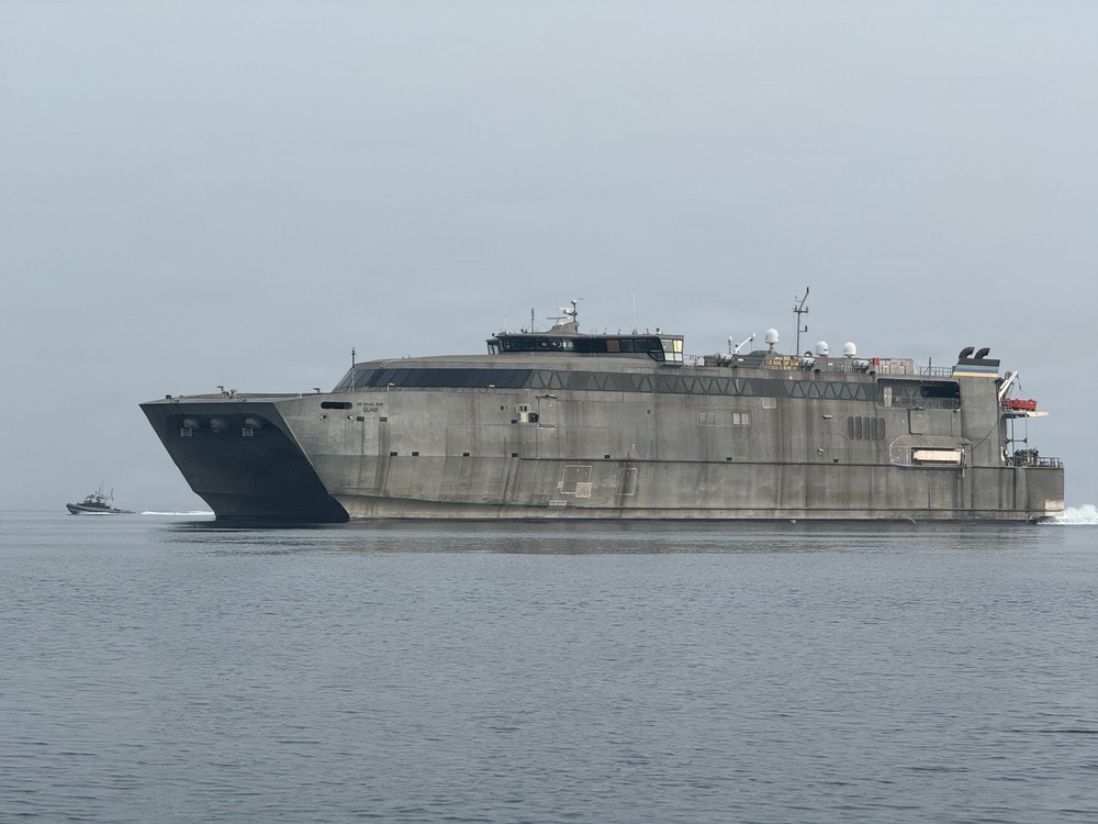 A large military catamaran vessel, consistent with a US Navy Expeditionary Fast Transport (EPF) ship, is underway on calm gray waters under an overcast sky. The vessel has a distinctive aluminum hull with a flat, wide superstructure and angular bow design. A smaller escort or patrol boat is visible in the background to the left.