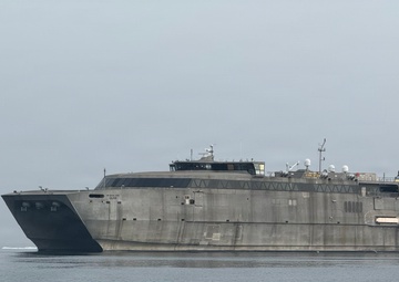 A large military catamaran vessel, consistent with a US Navy Expeditionary Fast Transport (EPF) ship, is underway on calm gray waters under an overcast sky. The vessel has a distinctive aluminum hull with a flat, wide superstructure and angular bow design. A smaller escort or patrol boat is visible in the background to the left.