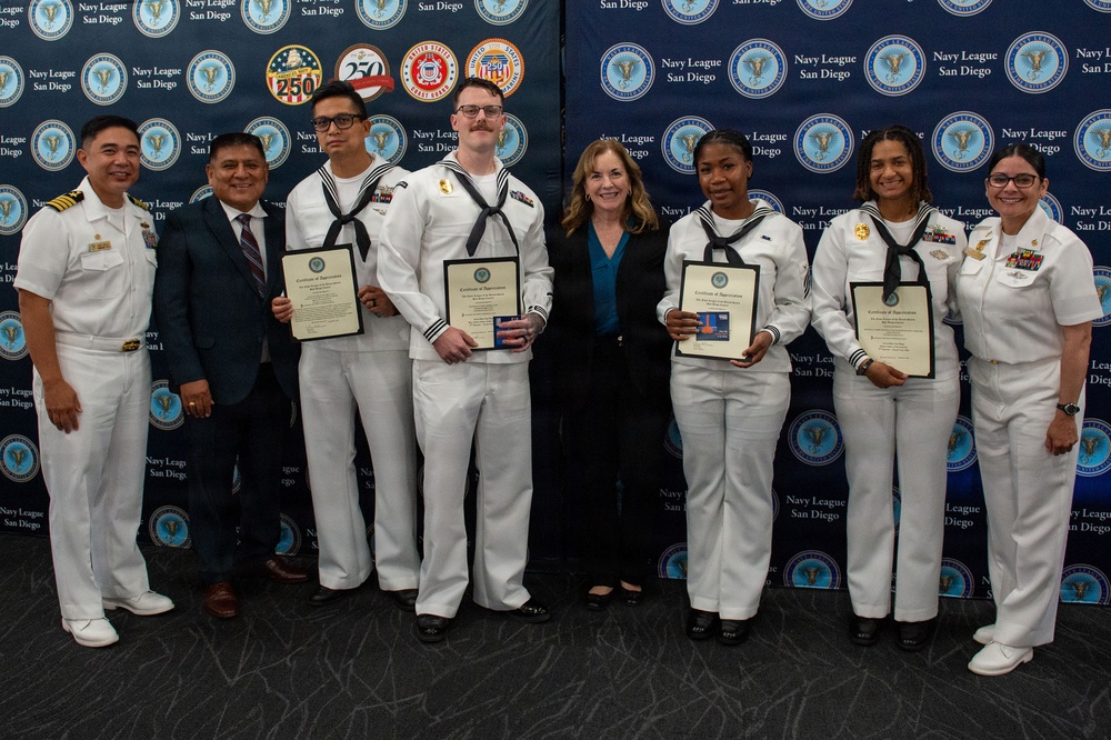 A group of eight individuals pose for a formal photo in front of a Navy League San Diego branded backdrop. Four individuals in U.S. Navy white dress uniforms are holding framed certificates of appreciation, flanked by officers and a civilian woman in business attire. The event appears to be an awards or recognition ceremony hosted by the Navy League San Diego.