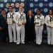 A group of eight individuals pose for a formal photo in front of a Navy League San Diego branded backdrop. Four individuals in U.S. Navy white dress uniforms are holding framed certificates of appreciation, flanked by officers and a civilian woman in business attire. The event appears to be an awards or recognition ceremony hosted by the Navy League San Diego.