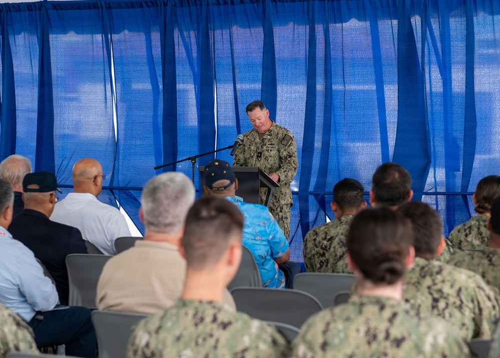 A U.S. Navy officer in digital camouflage (NWU Type III) uniform stands at a podium and speaks into a microphone before an audience of seated military personnel and civilians. The event appears to be a formal military ceremony or briefing held under a blue curtained backdrop, likely outdoors under a tent or canopy. The audience consists of a mix of uniformed Navy sailors and civilian attendees seated in folding chairs.