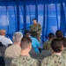 A U.S. Navy officer in digital camouflage (NWU Type III) uniform stands at a podium and speaks into a microphone before an audience of seated military personnel and civilians. The event appears to be a formal military ceremony or briefing held under a blue curtained backdrop, likely outdoors under a tent or canopy. The audience consists of a mix of uniformed Navy sailors and civilian attendees seated in folding chairs.