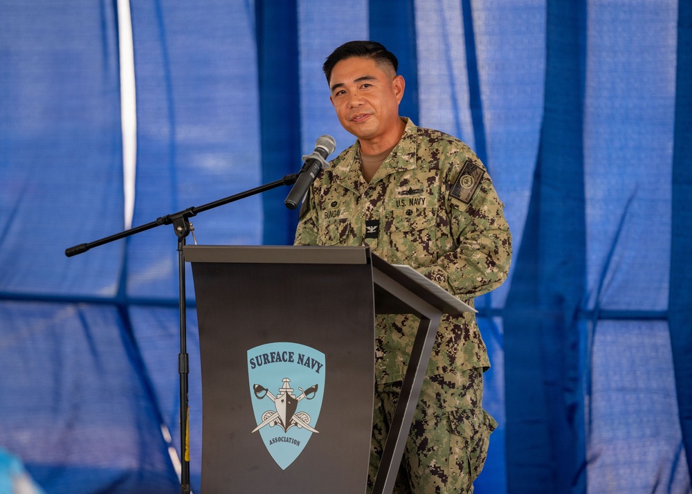 A U.S. Navy officer in NWU Type III (digital camouflage) uniform is speaking at a podium bearing the Surface Navy Association logo. The officer appears to be delivering a speech or remarks at an official military event held under a blue tent or canopy. A name tag is visible on the uniform, and the officer holds the rank of Captain (O-6) based on the eagle insignia visible on the collar.