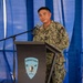 A U.S. Navy officer in NWU Type III (digital camouflage) uniform is speaking at a podium bearing the Surface Navy Association logo. The officer appears to be delivering a speech or remarks at an official military event held under a blue tent or canopy. A name tag is visible on the uniform, and the officer holds the rank of Captain (O-6) based on the eagle insignia visible on the collar.