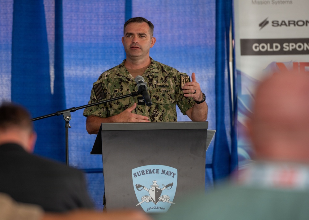 A U.S. Navy officer with the name tag 'Sheldon' and rank of Captain (O-6, indicated by eagle insignia) is speaking at a podium bearing the Surface Navy Association logo. He is wearing the Navy Working Uniform (NWU Type III) camouflage pattern and appears to be addressing an audience at what appears to be a sponsored event or symposium. A sponsor banner including 'SARON' and 'GOLD SPONSOR' text is visible in the background.