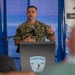 A U.S. Navy officer with the name tag 'Sheldon' and rank of Captain (O-6, indicated by eagle insignia) is speaking at a podium bearing the Surface Navy Association logo. He is wearing the Navy Working Uniform (NWU Type III) camouflage pattern and appears to be addressing an audience at what appears to be a sponsored event or symposium. A sponsor banner including 'SARON' and 'GOLD SPONSOR' text is visible in the background.