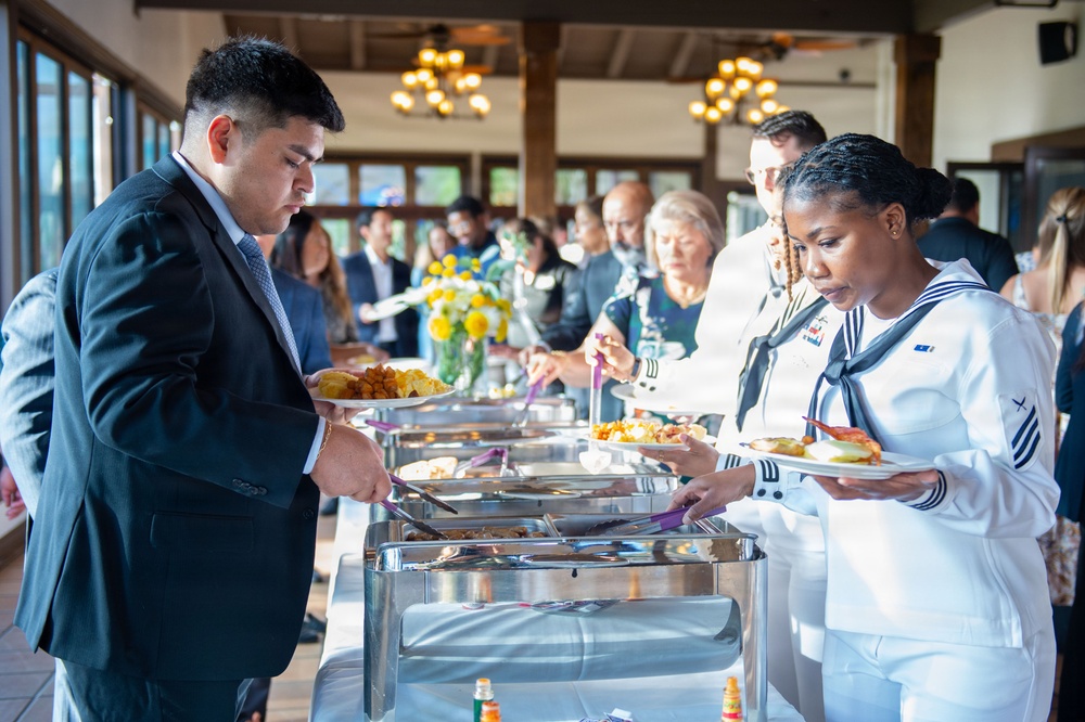 A formal buffet event is taking place in a well-lit banquet hall or restaurant setting. Attendees in business attire and at least one person in a U.S. Navy dress white uniform are serving themselves from a row of stainless steel chafing dishes. The background shows a crowd of additional guests mingling near floral centerpieces and chandeliers.