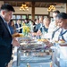 A formal buffet event is taking place in a well-lit banquet hall or restaurant setting. Attendees in business attire and at least one person in a U.S. Navy dress white uniform are serving themselves from a row of stainless steel chafing dishes. The background shows a crowd of additional guests mingling near floral centerpieces and chandeliers.