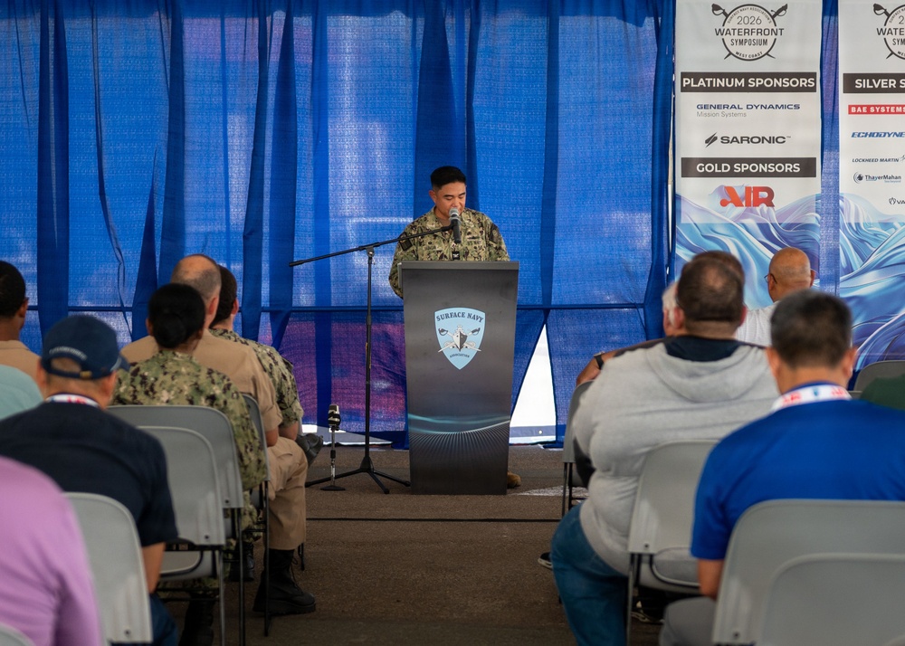 A military personnel in camouflage uniform is speaking at a podium bearing the 'Surface Navy Association' logo during the 2026 Waterfront Symposium West Coast event. An audience of mixed military and civilian attendees is seated in rows facing the speaker. Sponsor banners are visible in the background, including Platinum Sponsors General Dynamics Mission Systems and Saronic, and Gold Sponsor AIR.