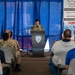 A military personnel in camouflage uniform is speaking at a podium bearing the 'Surface Navy Association' logo during the 2026 Waterfront Symposium West Coast event. An audience of mixed military and civilian attendees is seated in rows facing the speaker. Sponsor banners are visible in the background, including Platinum Sponsors General Dynamics Mission Systems and Saronic, and Gold Sponsor AIR.