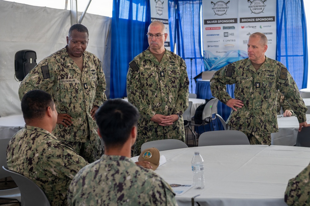 A group of U.S. Navy personnel in NWU Type III (digital camouflage) uniforms are gathered under a tent at what appears to be a 2024 Waterfront Symposium event. Three standing officers, identified by name tapes as Westmoreland, Lynch, and Schmidt, appear to be addressing or briefing seated sailors at a round table. The setting includes sponsor banners, blue curtains, and round tables with white tablecloths.