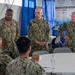 A group of U.S. Navy personnel in NWU Type III (digital camouflage) uniforms are gathered under a tent at what appears to be a 2024 Waterfront Symposium event. Three standing officers, identified by name tapes as Westmoreland, Lynch, and Schmidt, appear to be addressing or briefing seated sailors at a round table. The setting includes sponsor banners, blue curtains, and round tables with white tablecloths.