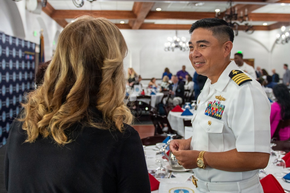 A U.S. Navy officer in white dress uniform is engaged in conversation with a civilian woman at what appears to be a formal military banquet or luncheon event. The officer displays multiple service ribbons, a gold warfare device, and rank insignia consistent with a senior officer (Captain or Commander). The setting is an elegant banquet hall with white-draped tables and patriotic red, white, and blue decorations.