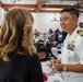 A U.S. Navy officer in white dress uniform is engaged in conversation with a civilian woman at what appears to be a formal military banquet or luncheon event. The officer displays multiple service ribbons, a gold warfare device, and rank insignia consistent with a senior officer (Captain or Commander). The setting is an elegant banquet hall with white-draped tables and patriotic red, white, and blue decorations.