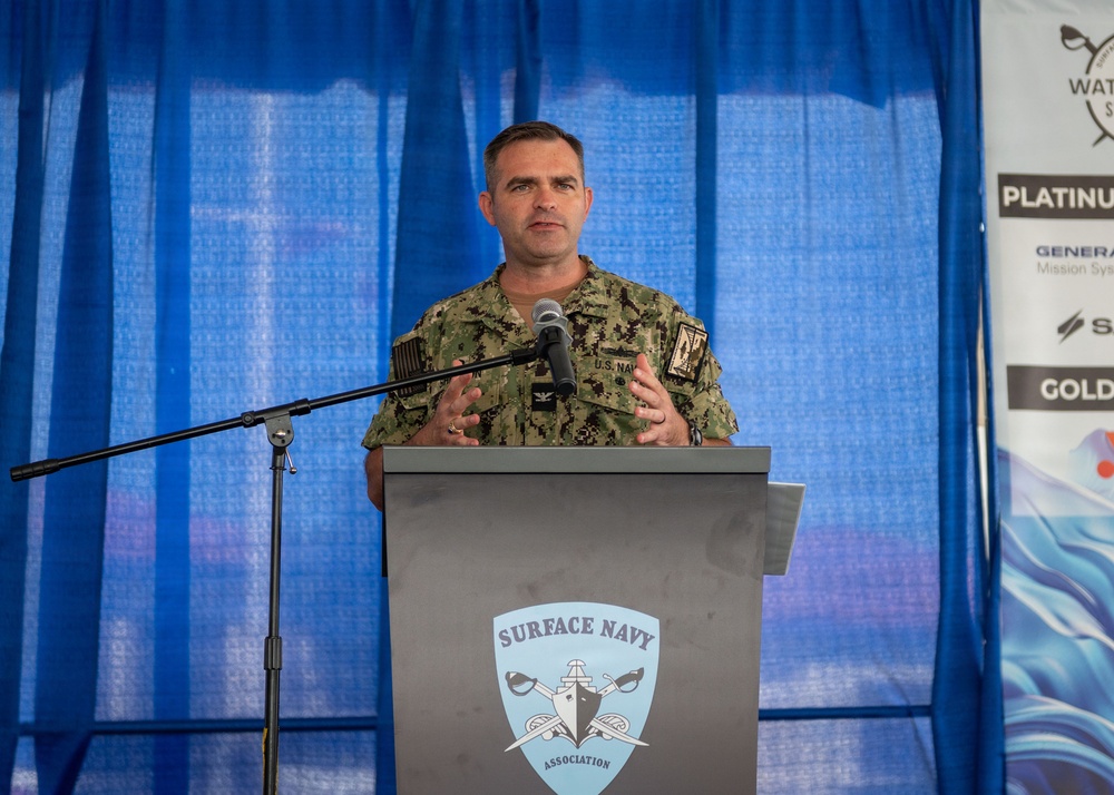 A U.S. Navy officer in digital camouflage uniform is speaking at a podium bearing the Surface Navy Association logo. The speaker appears to be addressing an audience at what appears to be a Surface Navy Association event, with sponsor banners visible in the background including 'Platinum' and 'Gold' level sponsors. The setting appears to be an outdoor or tent venue with blue curtain backdrop.