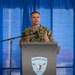 A U.S. Navy officer in digital camouflage uniform is speaking at a podium bearing the Surface Navy Association logo. The speaker appears to be addressing an audience at what appears to be a Surface Navy Association event, with sponsor banners visible in the background including 'Platinum' and 'Gold' level sponsors. The setting appears to be an outdoor or tent venue with blue curtain backdrop.