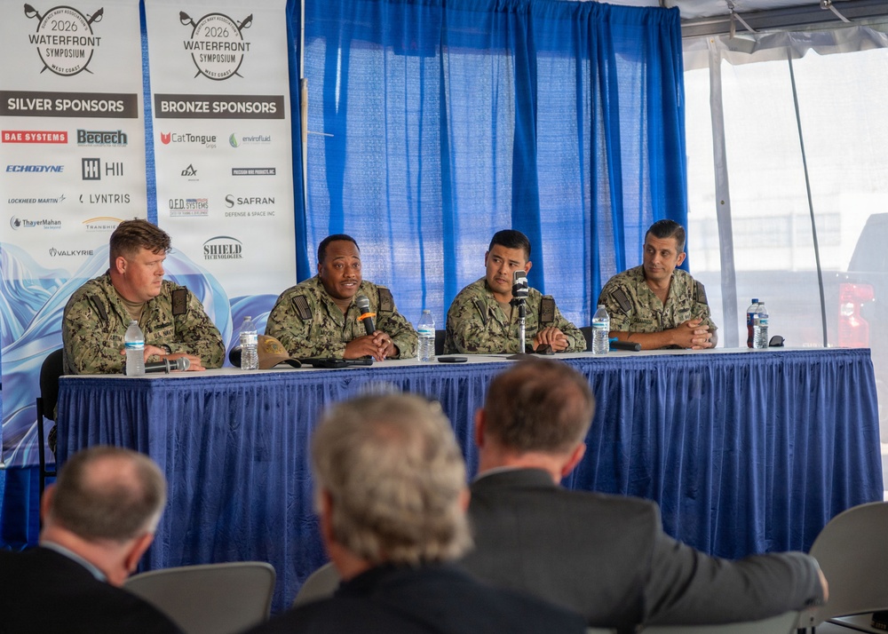 Four U.S. Navy personnel in camouflage uniforms are seated at a draped panel table during the 2026 Waterfront Symposium West Coast event. One panelist appears to be speaking into a microphone while the others listen. An audience is visible from behind in the foreground, facing the panel.