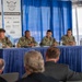 Four U.S. Navy personnel in camouflage uniforms are seated at a draped panel table during the 2026 Waterfront Symposium West Coast event. One panelist appears to be speaking into a microphone while the others listen. An audience is visible from behind in the foreground, facing the panel.