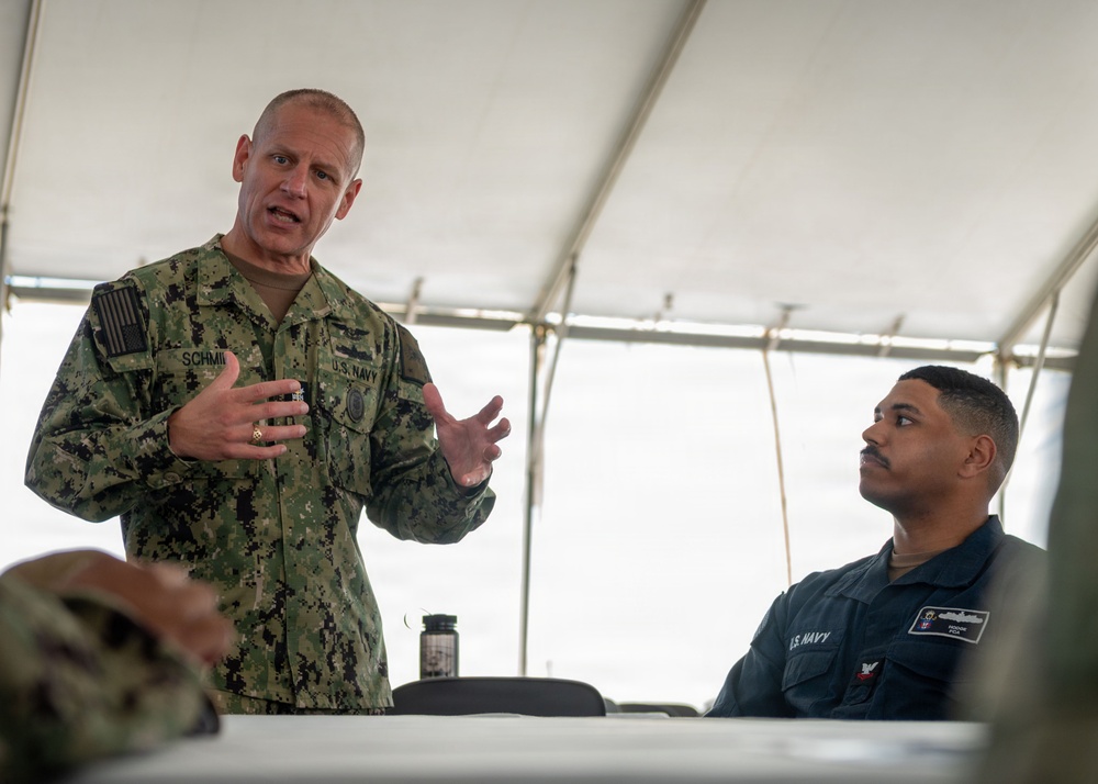 A U.S. Navy officer with the name tape 'SCHMI_' (partially visible) wearing NWU Type III camouflage uniform is speaking and gesturing with both hands under a white tent. A second U.S. Navy sailor in dark blue NWU Type I uniform is seated at a table listening attentively. The setting appears to be an outdoor briefing or meeting event.