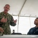 A U.S. Navy officer with the name tape 'SCHMI_' (partially visible) wearing NWU Type III camouflage uniform is speaking and gesturing with both hands under a white tent. A second U.S. Navy sailor in dark blue NWU Type I uniform is seated at a table listening attentively. The setting appears to be an outdoor briefing or meeting event.