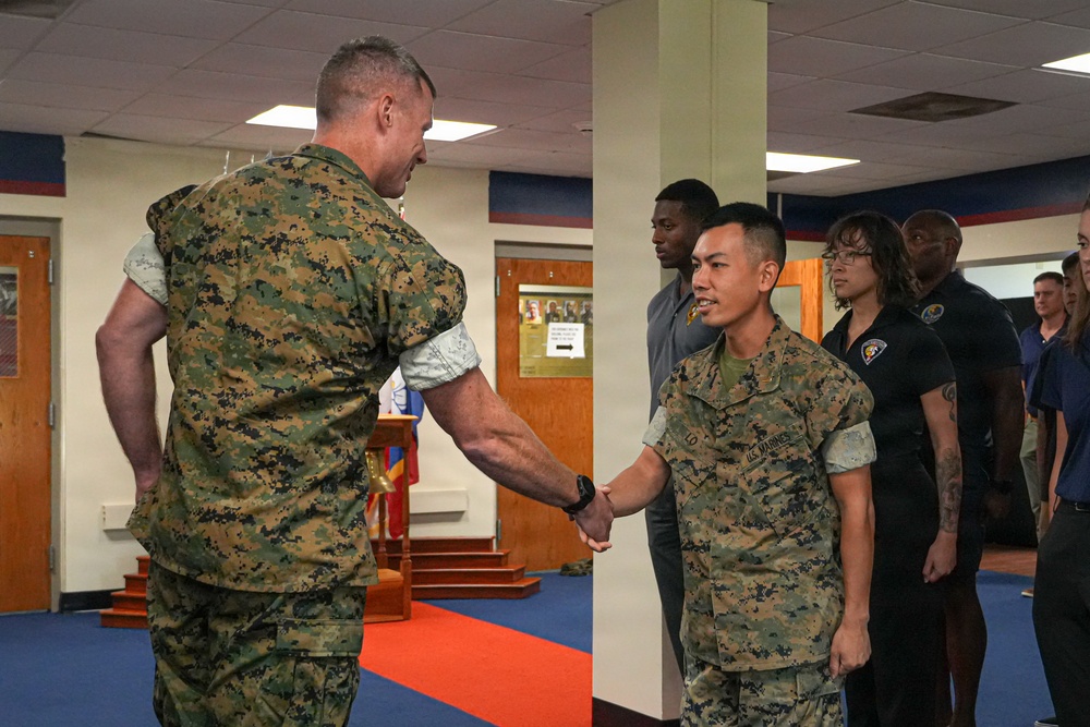 Two U.S. Marines in woodland MARPAT camouflage uniforms are shaking hands in what appears to be an indoor military facility with a matted floor. The scene suggests a formal or ceremonial interaction, possibly a recognition, graduation, or greeting event. Several civilians and other personnel are visible standing in the background observing the exchange.