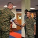 Two U.S. Marines in woodland MARPAT camouflage uniforms are shaking hands in what appears to be an indoor military facility with a matted floor. The scene suggests a formal or ceremonial interaction, possibly a recognition, graduation, or greeting event. Several civilians and other personnel are visible standing in the background observing the exchange.
