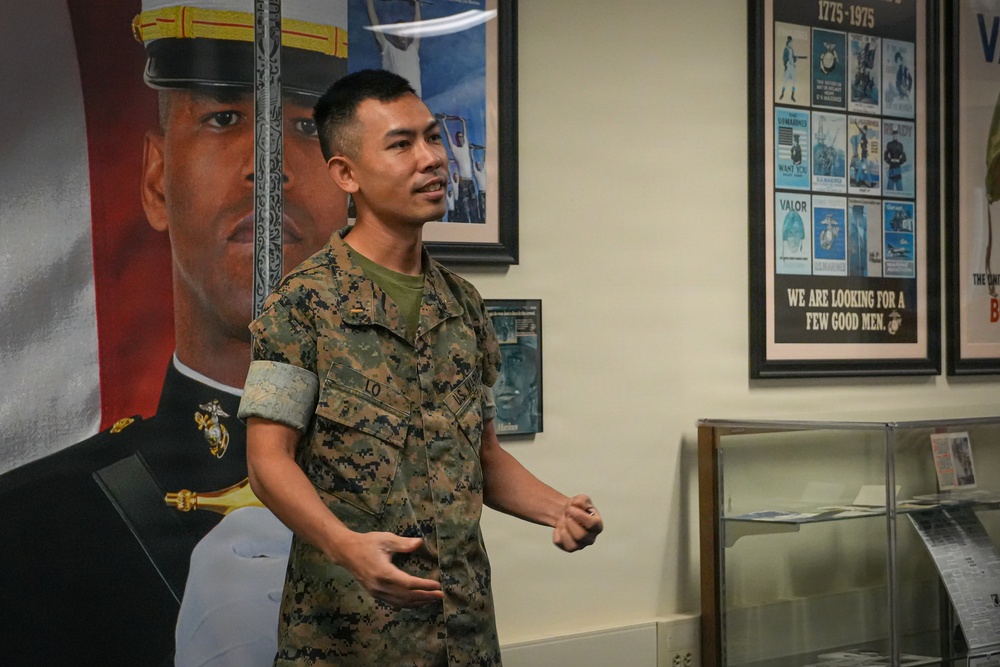 A U.S. Marine in woodland MARPAT camouflage uniform is speaking or presenting inside what appears to be a military recruiting office or museum setting. The Marine, whose name tape reads 'LO', is gesturing with his hands while standing in front of recruitment posters and display cases. The background features historical U.S. Marine Corps recruitment materials spanning 1775–1975.
