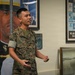 A U.S. Marine in woodland MARPAT camouflage uniform is speaking or presenting inside what appears to be a military recruiting office or museum setting. The Marine, whose name tape reads 'LO', is gesturing with his hands while standing in front of recruitment posters and display cases. The background features historical U.S. Marine Corps recruitment materials spanning 1775–1975.