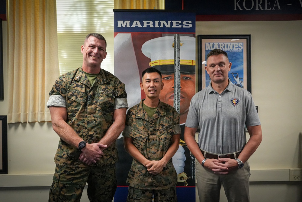 Three individuals pose for a formal photo in what appears to be a U.S. Marine Corps recruiting or administrative office. Two of the individuals are wearing U.S. Marine Corps camouflage uniforms, while the third is in civilian attire wearing a polo shirt with a military insignia. A Marines recruitment banner and framed Marines poster are visible in the background.