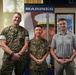 Three individuals pose for a formal photo in what appears to be a U.S. Marine Corps recruiting or administrative office. Two of the individuals are wearing U.S. Marine Corps camouflage uniforms, while the third is in civilian attire wearing a polo shirt with a military insignia. A Marines recruitment banner and framed Marines poster are visible in the background.