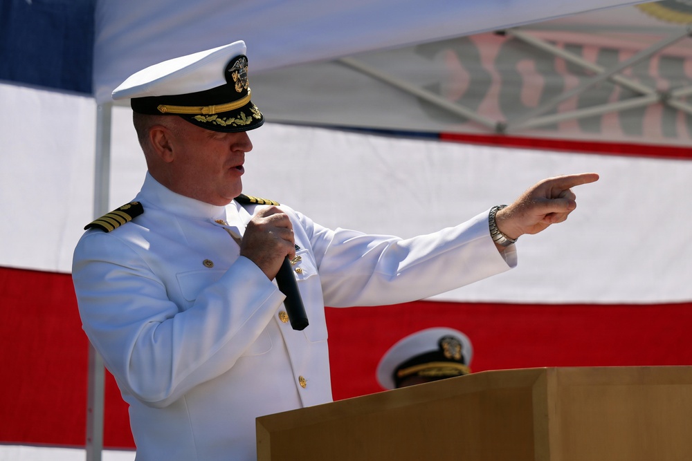A senior U.S. Navy officer in white dress uniform is speaking at a podium while holding a microphone and pointing with his right hand. The event appears to be a formal military ceremony, likely a change of command or commissioning, held outdoors with an American flag prominently displayed in the background. A second naval officer is partially visible behind the podium.