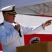 A senior U.S. Navy officer in white dress uniform is speaking at a podium while holding a microphone and pointing with his right hand. The event appears to be a formal military ceremony, likely a change of command or commissioning, held outdoors with an American flag prominently displayed in the background. A second naval officer is partially visible behind the podium.
