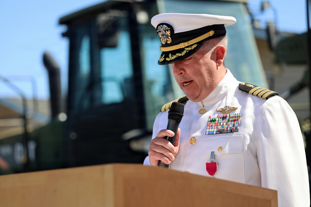 A senior U.S. Navy officer in white dress uniform is speaking at a podium while holding a microphone. He is wearing a combination cover with gold oak leaf scrambled eggs indicating flag or senior officer rank, along with multiple service ribbons and medals. The setting appears to be an outdoor ceremony with construction equipment visible in the background.