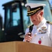 A senior U.S. Navy officer in white dress uniform is speaking at a podium while holding a microphone. He is wearing a combination cover with gold oak leaf scrambled eggs indicating flag or senior officer rank, along with multiple service ribbons and medals. The setting appears to be an outdoor ceremony with construction equipment visible in the background.