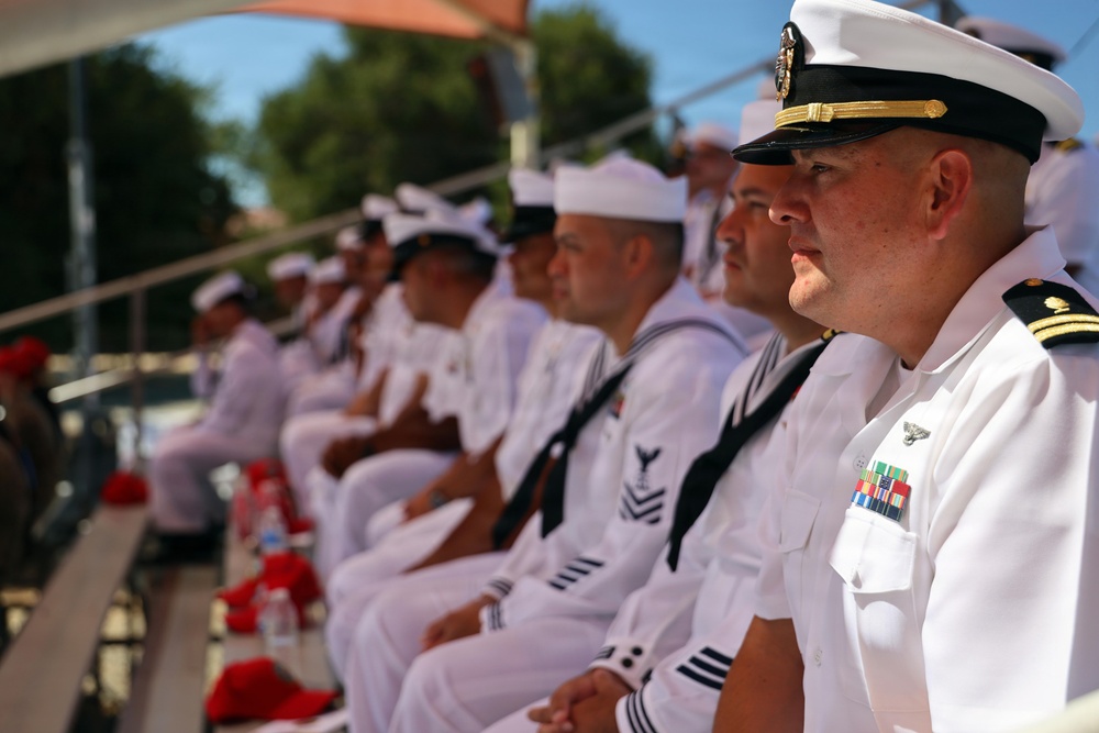 A group of naval personnel dressed in white dress uniforms are seated in rows, appearing to attend a formal military ceremony or event outdoors. The foreground figure is a senior officer wearing a combination cover (peaked cap) with gold braid and a ribbon rack with multiple service medals. The setting appears to be an outdoor venue with trees and blue sky visible in the background.
