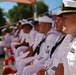 A group of naval personnel dressed in white dress uniforms are seated in rows, appearing to attend a formal military ceremony or event outdoors. The foreground figure is a senior officer wearing a combination cover (peaked cap) with gold braid and a ribbon rack with multiple service medals. The setting appears to be an outdoor venue with trees and blue sky visible in the background.