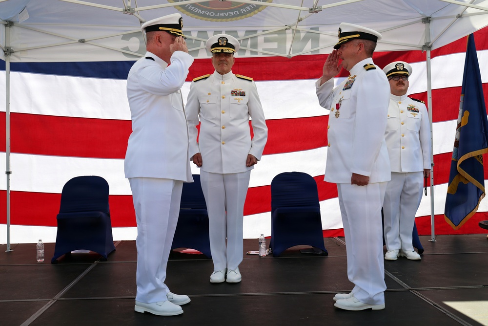 A formal U.S. Navy change of command ceremony is taking place on an outdoor stage. Two naval officers in white dress uniforms are exchanging salutes in the foreground, while two additional officers stand at attention in the background. A large American flag serves as a backdrop, along with what appears to be a Navy unit flag on the right side.