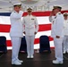 A formal U.S. Navy change of command ceremony is taking place on an outdoor stage. Two naval officers in white dress uniforms are exchanging salutes in the foreground, while two additional officers stand at attention in the background. A large American flag serves as a backdrop, along with what appears to be a Navy unit flag on the right side.