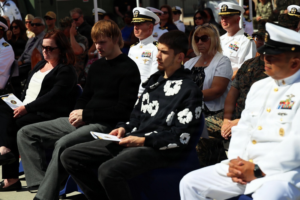 A formal military ceremony with a mixed audience of uniformed naval/military personnel and civilians seated in rows. The attendees appear solemn and attentive, suggesting a significant official event such as a commissioning, retirement, or memorial ceremony. Several Navy officers in white dress uniforms are visible alongside civilians in casual and semi-formal attire.
