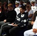 A formal military ceremony with a mixed audience of uniformed naval/military personnel and civilians seated in rows. The attendees appear solemn and attentive, suggesting a significant official event such as a commissioning, retirement, or memorial ceremony. Several Navy officers in white dress uniforms are visible alongside civilians in casual and semi-formal attire.