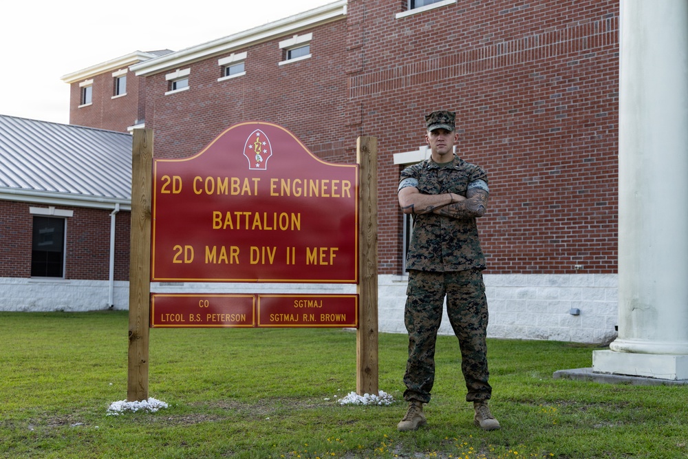 A U.S. Marine in digital camouflage (MARPAT) uniform stands with arms crossed beside the unit sign for the 2nd Combat Engineer Battalion, 2nd Marine Division, II Marine Expeditionary Force. The sign lists the Commanding Officer as LTCOL B.S. Peterson and the Sergeant Major as SGTMAJ R.N. Brown. The setting appears to be a military installation with a brick building in the background.