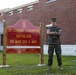 A U.S. Marine in digital camouflage (MARPAT) uniform stands with arms crossed beside the unit sign for the 2nd Combat Engineer Battalion, 2nd Marine Division, II Marine Expeditionary Force. The sign lists the Commanding Officer as LTCOL B.S. Peterson and the Sergeant Major as SGTMAJ R.N. Brown. The setting appears to be a military installation with a brick building in the background.